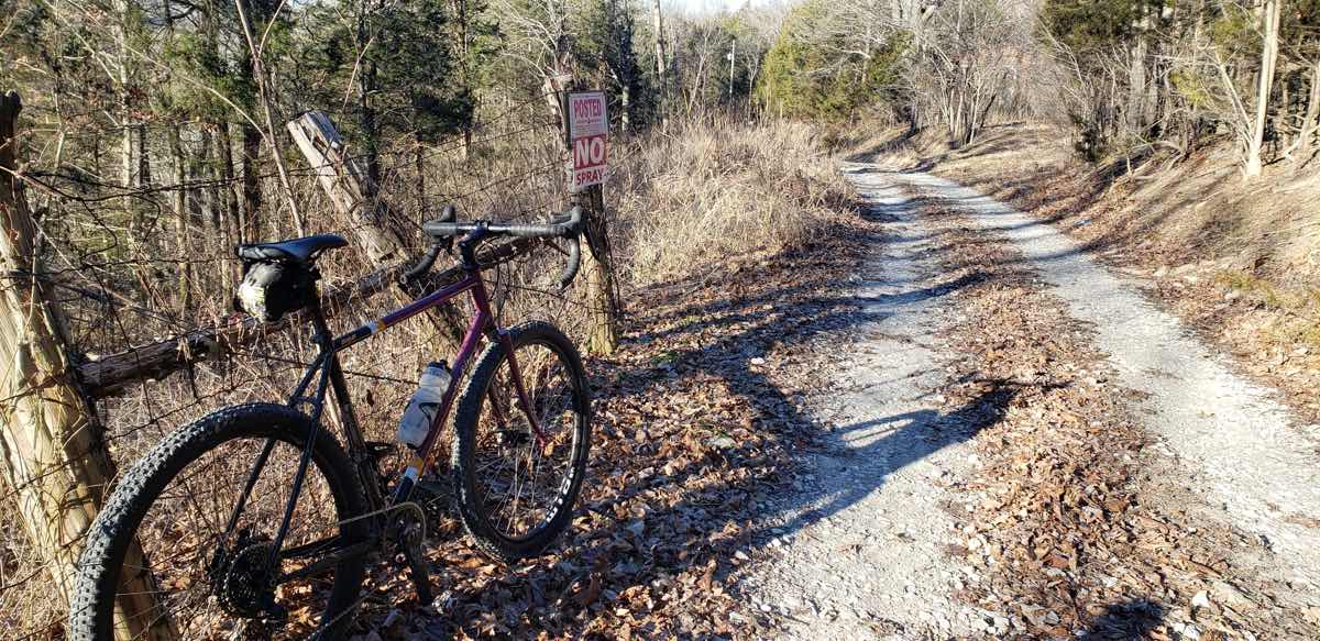 bikerumor pic of the day a gravel bicycle leans against a wood and barbed wire fence on the side of a gravel road with scrub brush surrounding, the sun is low in the sky and shadows are long.