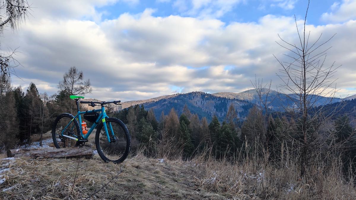 a bicycle is on a grassy clearing near a stand of trees on the top of a mountain overlooking a rolling mountain range, there are clouds in the sky with small patches of blue peeking out.