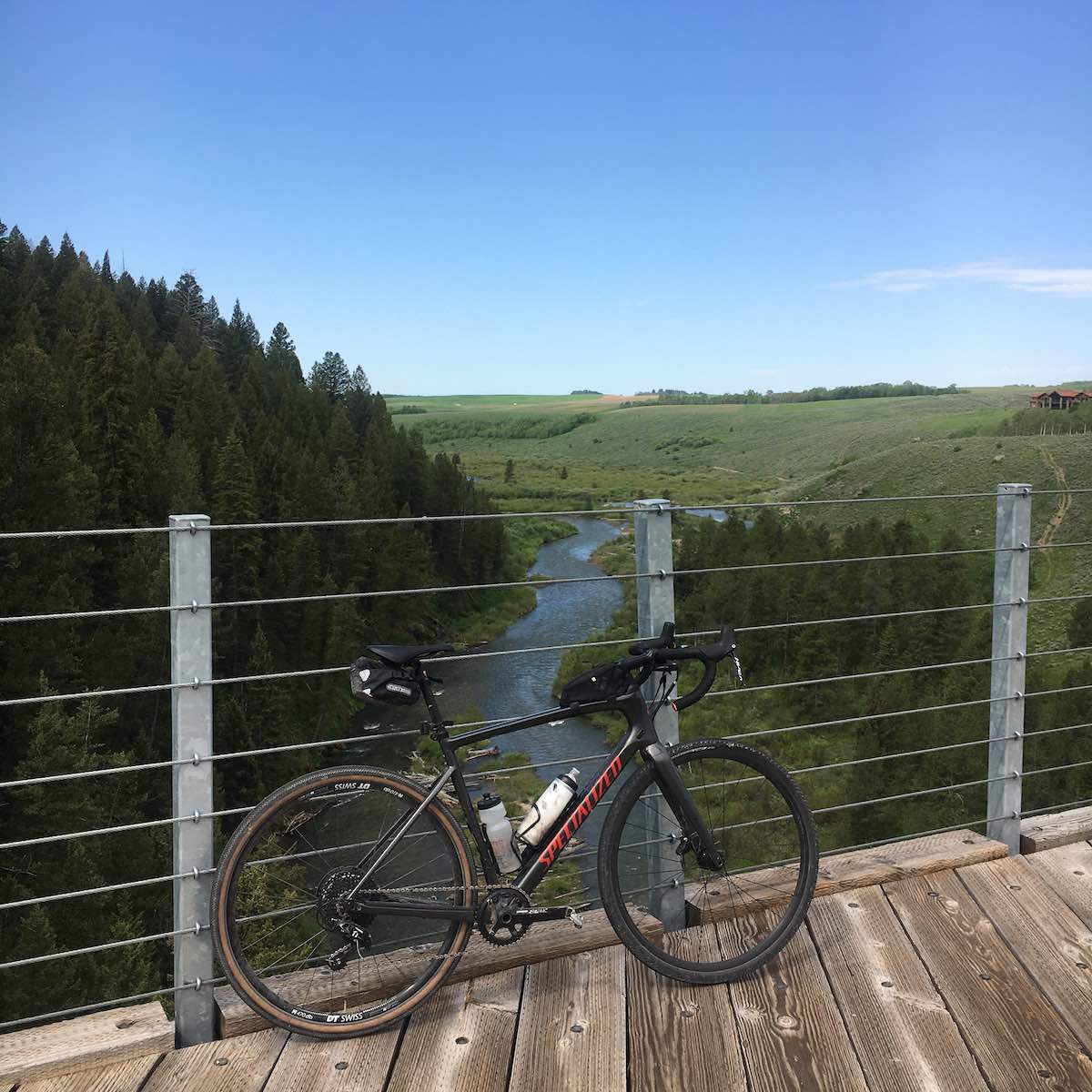 bikerumor pic of the day a bicycle leans against the wire railing of a wood bridge overlooking a stream among green pines trees and green grassy hill, the sky is clear and blue