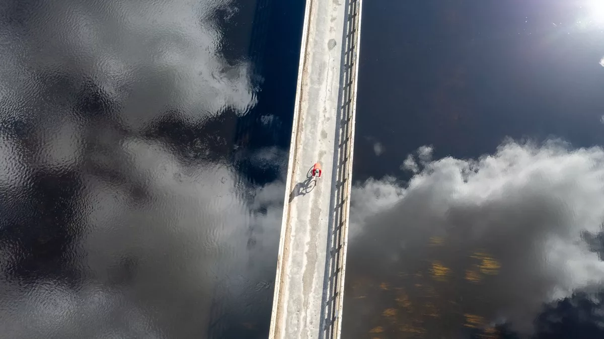 bikerumor pic of the day a cyclist crosses a bridge in an aril view looking down as the bridge crosses a clear calm lake reflecting the sky