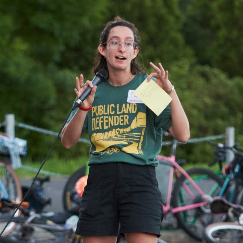 A woman speaking into a microphone wearing the Public Land Defender t-shirt